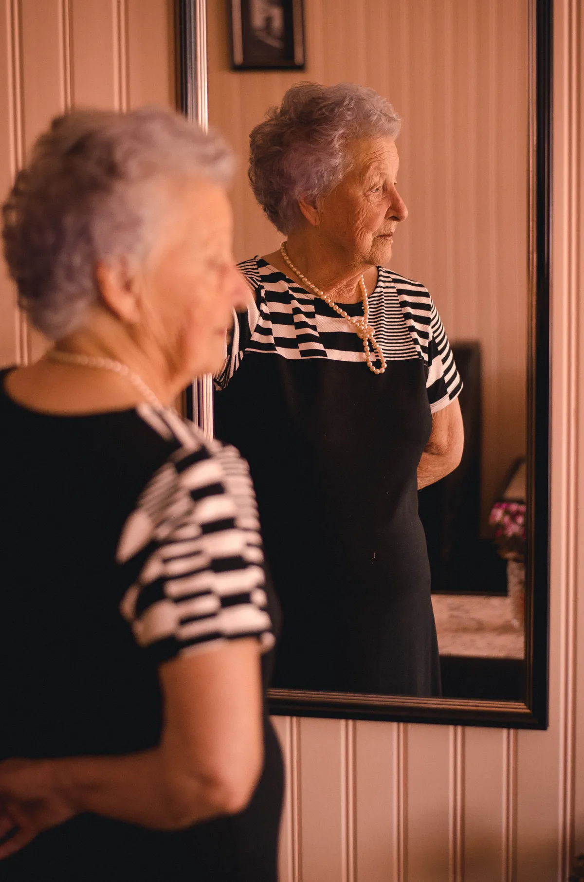 Anciana parada frente a un espejo mirando hacia un lado. | Foto: Pexels 