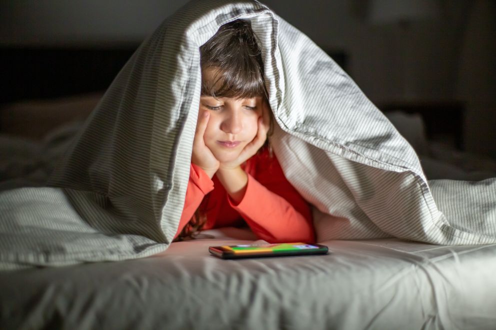 Un niño recostado en una cama bajo una manta mirando un teléfono celular. | Foto: Pexels