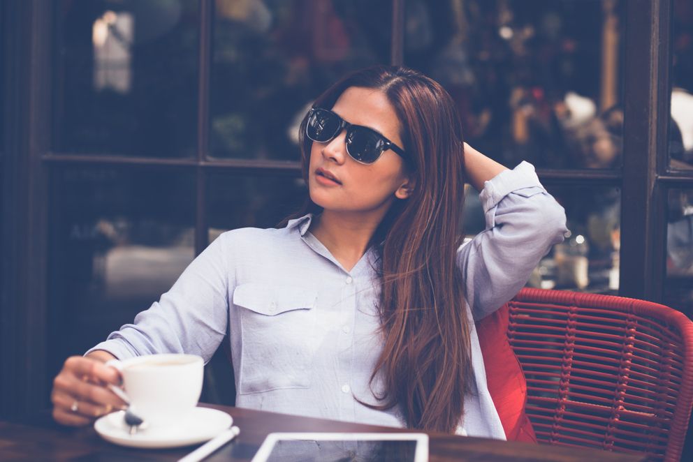 Una joven con gafas de sol sentada frente a una mesa bebiendo un café. | Foto: Pexels