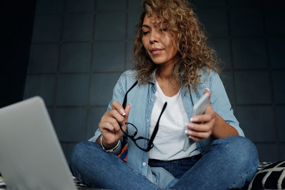 Una mujer sentada con su teléfono celular en su mano, mirando una computadora portátil. | Foto: Pexels