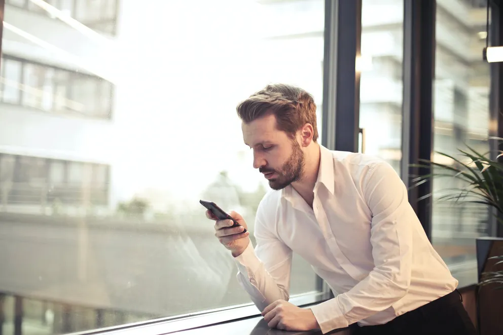 Hombre mirando un teléfono celular frente a un ventanal. | Foto: Pexels