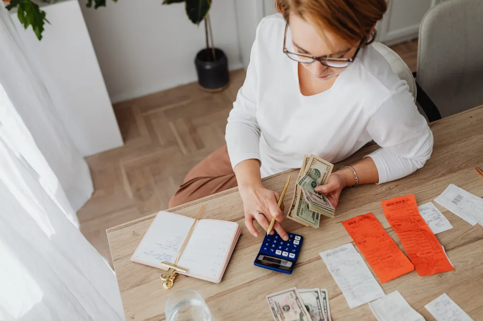Una mujer sentada frente a una mesa con una calculadora, facturas y unos billetes en su mano. | Foto: Pexels