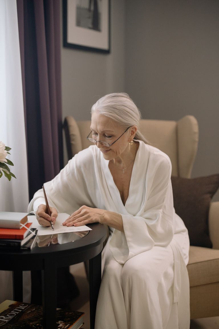 Una mujer mayor sentada junto a una mesa escribiendo en una hoja de papel. | Foto: Pexels