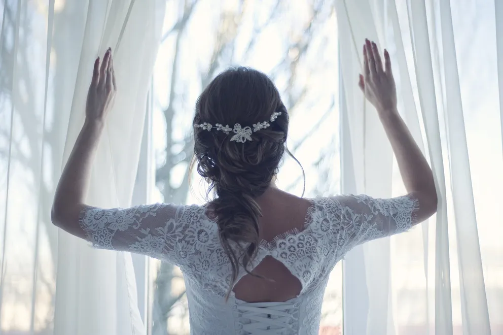 Une mariée dans une robe de mariée en dentelle debout près d'une grande fenêtre. | Source : Pixabay