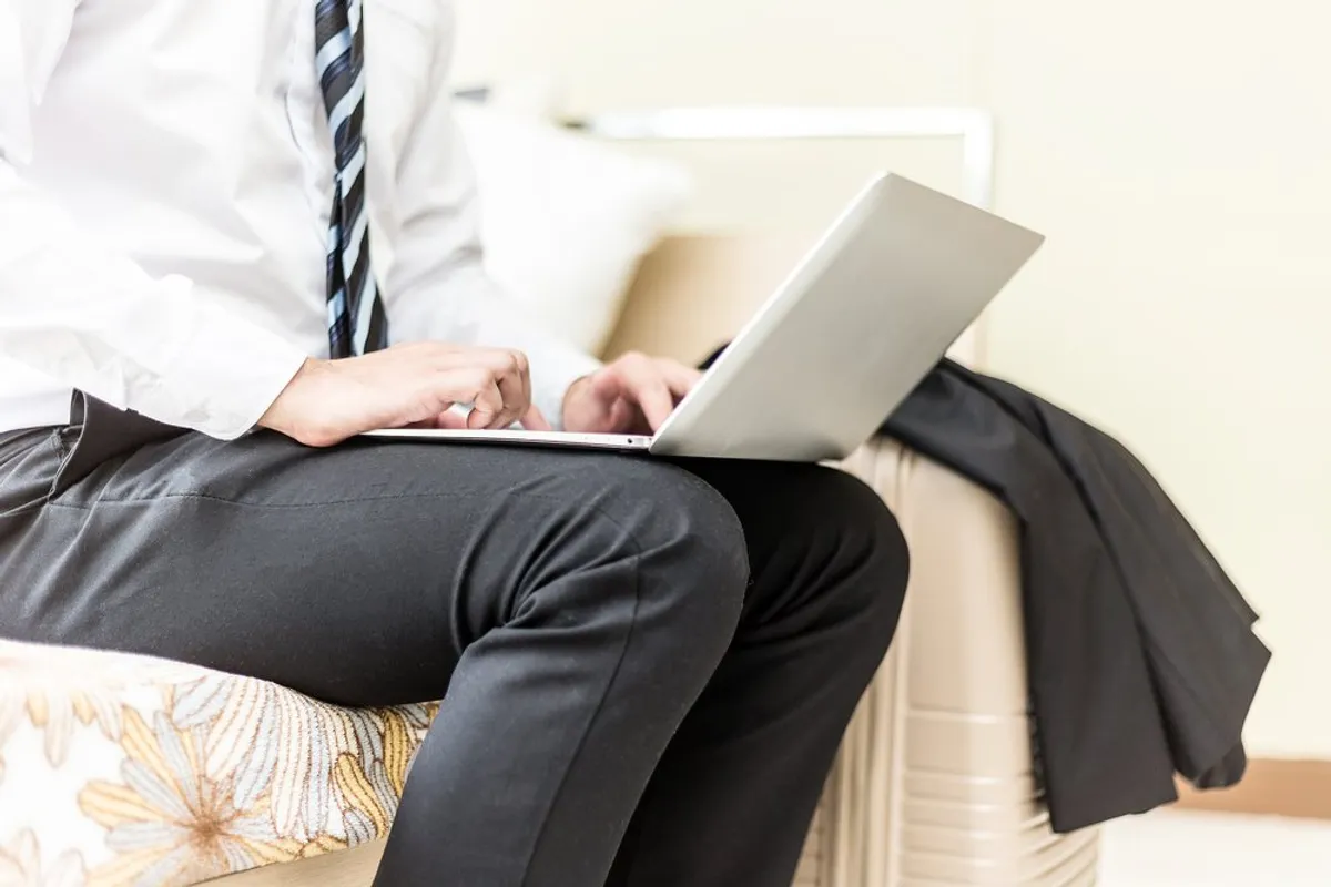 A businessman working on a laptop while sitting on a bed with his travel bag and jacket ready | Photo: Shutterstock/Yuttana Jaowattana