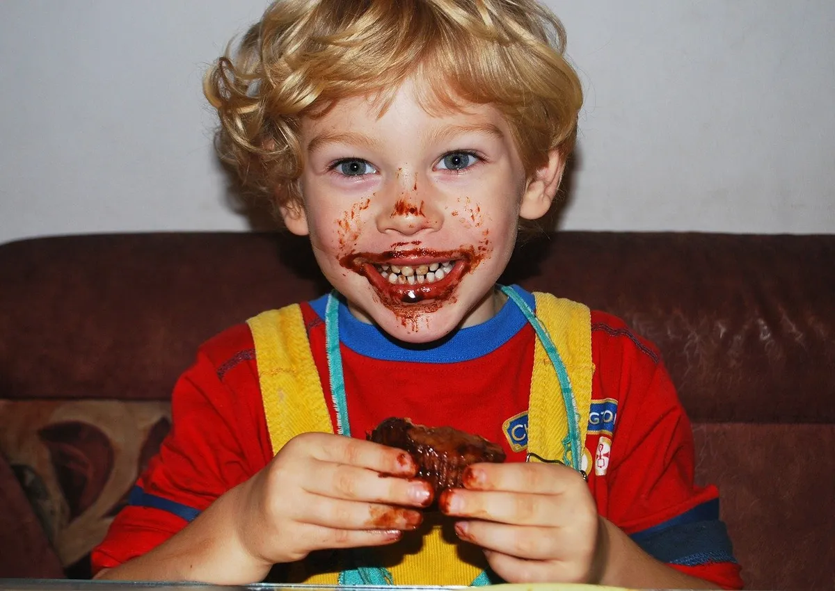 Ein kleiner Junge lächelt breit, während sein Mund mit dem Saft des Fleisches verschmiert ist, das er gerade isst | Quelle: Pixabay/Mojpe