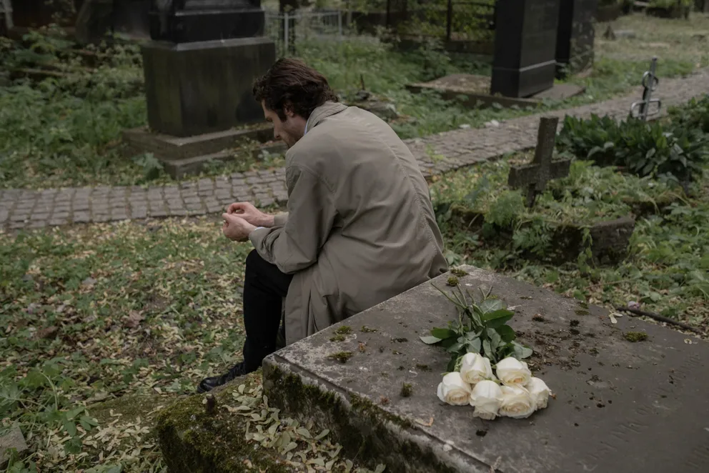 Un hombre afligido sentado junto a una tumba con flores. | Foto: Pexels
